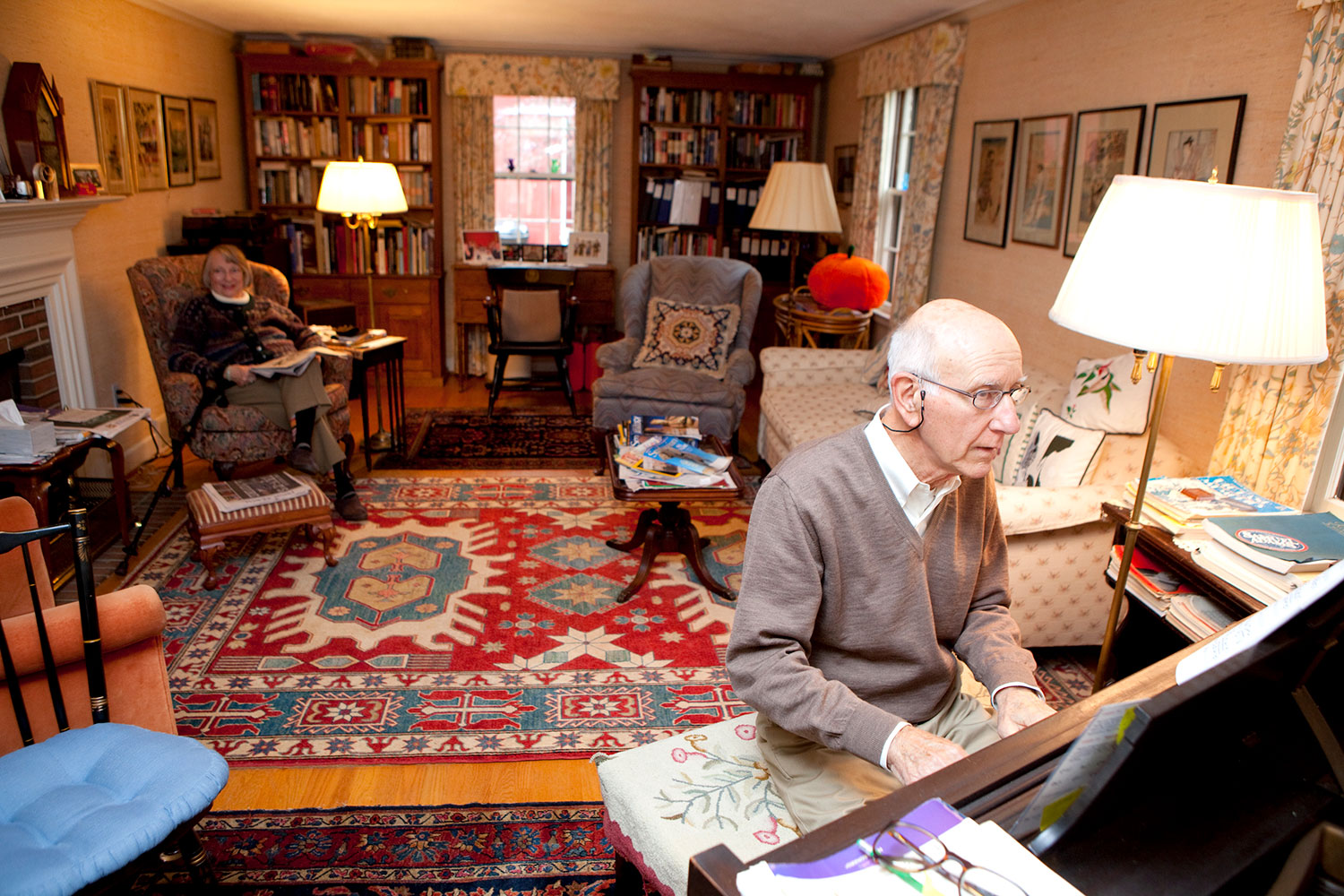 Man plays piano in living room while woman seated in a chair looks on.