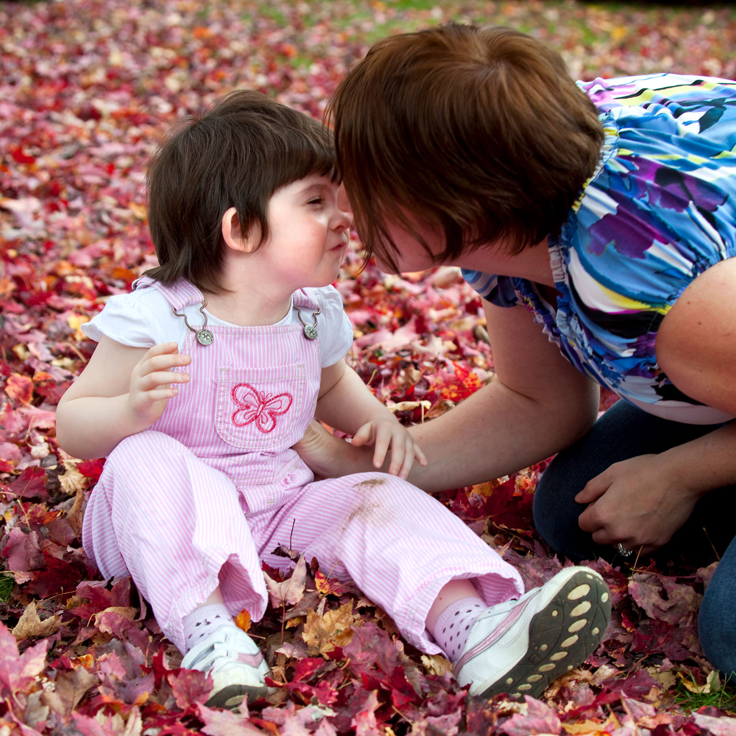 Toddler seated in autumn leaves outside. Adult kneels beside her. They give each other a kiss.